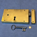 A brass door lockset with a rectangular lock plate, matching latch bolt, and an antique-style key, displayed on a textured blue background with a coin for scale.