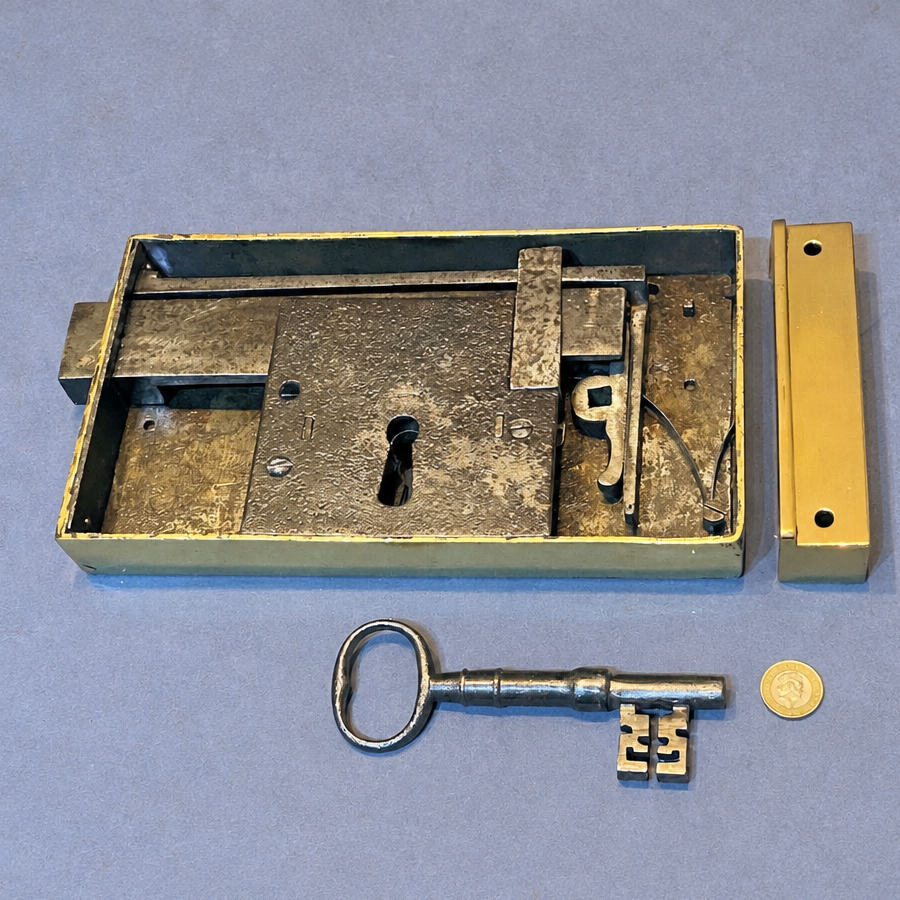 An antique metal lock mechanism set with a brass-toned rectangular lock body, a matching vertical plate, a silver key, and a small coin for scale, displayed on a blue-gray background.