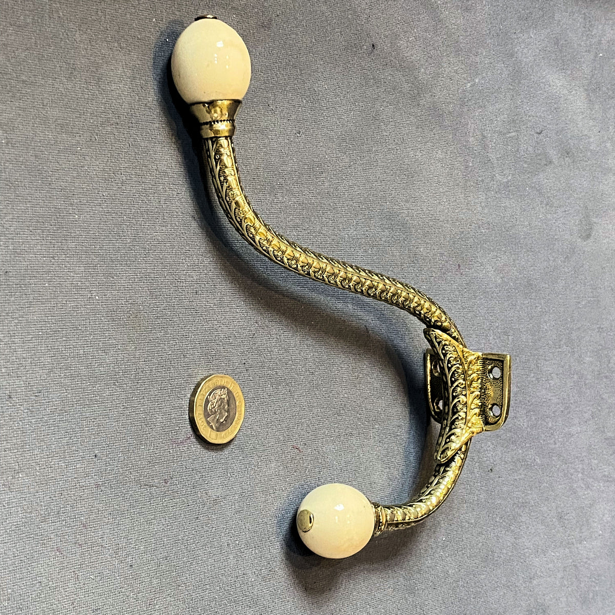 An antique brass S-hook wall hook with two cream-colored ceramic or ivory ball finials, displayed on a gray textured background next to a coin for scale.