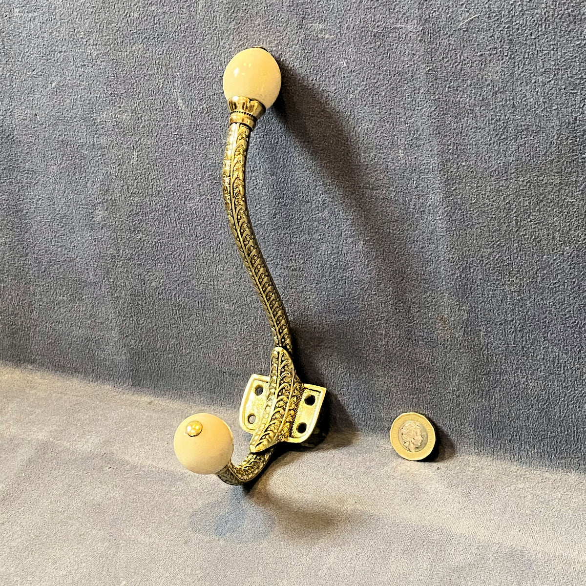 An ornate vintage-style coat rack hook with a textured gold-toned metal stem and two round cream-colored end knobs, shown against a gray fabric backdrop with a coin for scale.